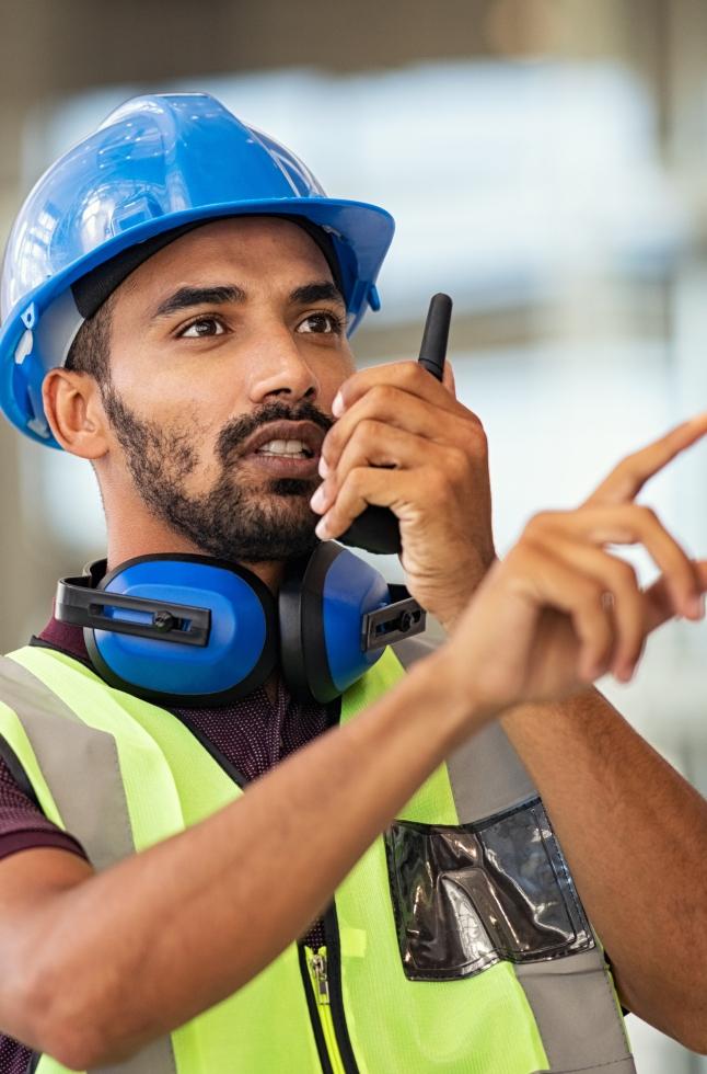 Komunikasi real-time untuk pengiriman dan armada logistik. Petugas logistik dengan rompi dan helm keselamatan sedang menggunakan radio PTT di area pergudangan.